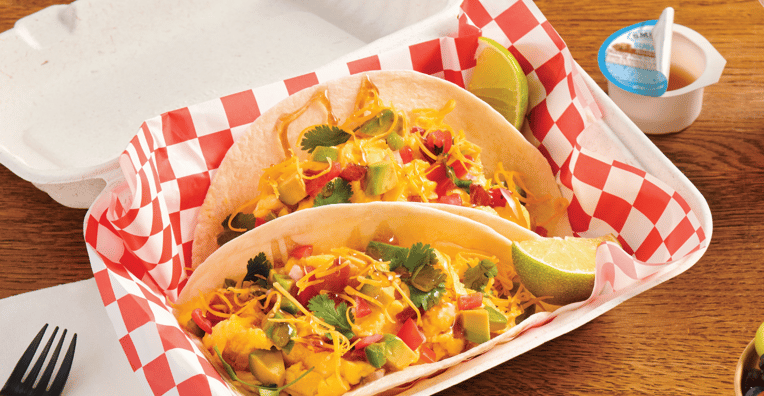 Two soft tacos filled with scrambled eggs, cheese, chopped tomatoes, cilantro, and diced vegetables, served in a checkered paper-lined takeout box with lime wedges and a side of maple syrup.