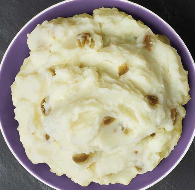 A bowl of creamy mashed potatoes with visible potato skin pieces, served in a purple bowl on a dark surface.