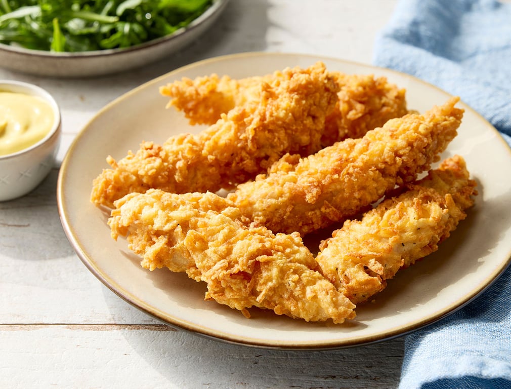 A plate of crispy golden chicken tenders sits on a light-colored table, with a small bowl of dipping sauce and a plate of leafy greens in the background. A blue napkin is partially visible to the side.