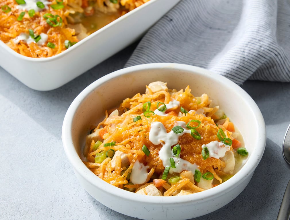 A white bowl filled with a cheesy casserole topped with chopped green onions and drizzled with a white sauce, sitting on a light gray surface next to a striped napkin.