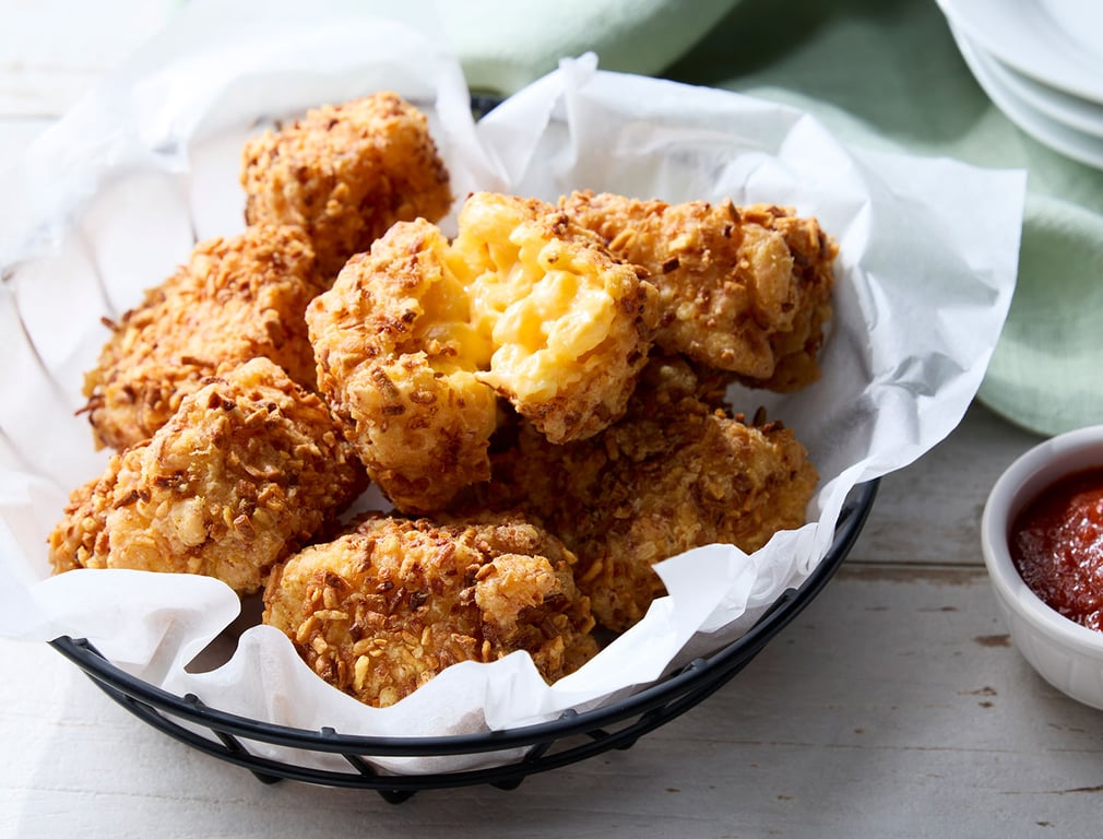 A basket lined with white parchment paper holds several pieces of crispy, golden-brown fried macaroni and cheese bites, with one piece broken open to show the creamy macaroni filling. A small dish of red dipping sauce is beside the basket.