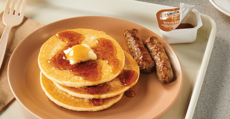 Three pancakes with syrup and butter, two sausage links, and a cup of breakfast syrup are served on a beige plate with a plastic fork and napkin on a cafeteria tray.