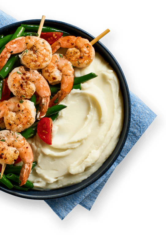 A black bowl with creamy mashed potatoes, grilled shrimp skewers, green beans, and red bell peppers, placed on a blue napkin.