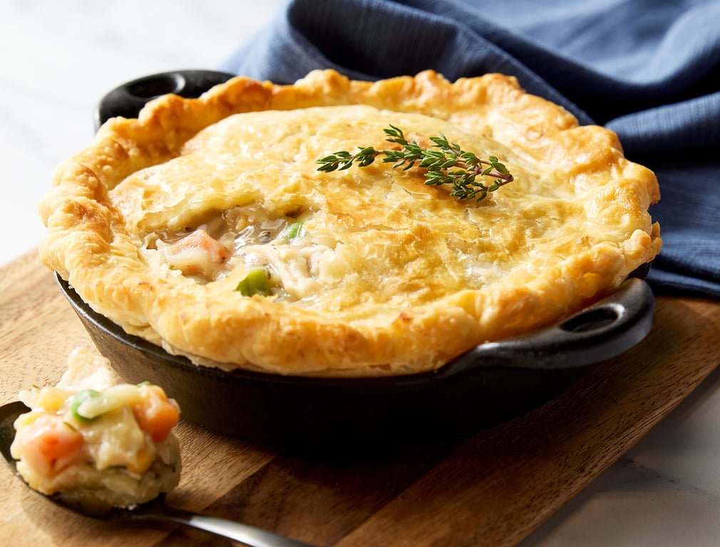 A golden, flaky pot pie sits in a black dish on a wooden board, topped with fresh thyme. A spoonful of creamy filling with vegetables rests beside it. A blue cloth is in the background.