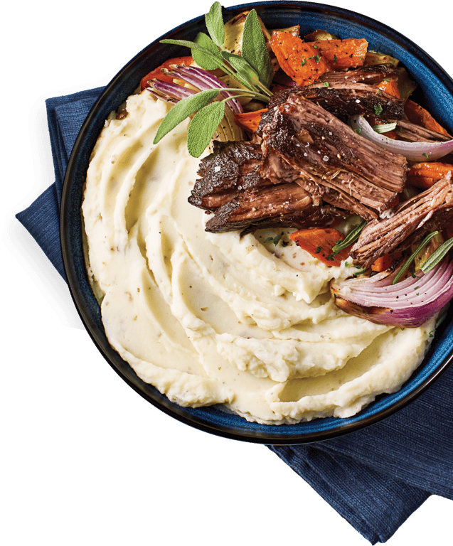 A plate of mashed potatoes topped with shredded pot roast, roasted carrots, red onions, and fresh herbs, set on a blue napkin with a fork beside it.
