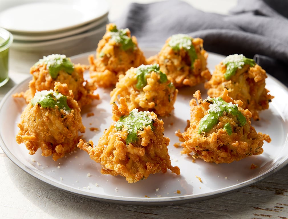 Eight crispy fritters topped with green sauce and a sprinkle of cheese are arranged on a white plate, with extra plates and a gray napkin in the background.