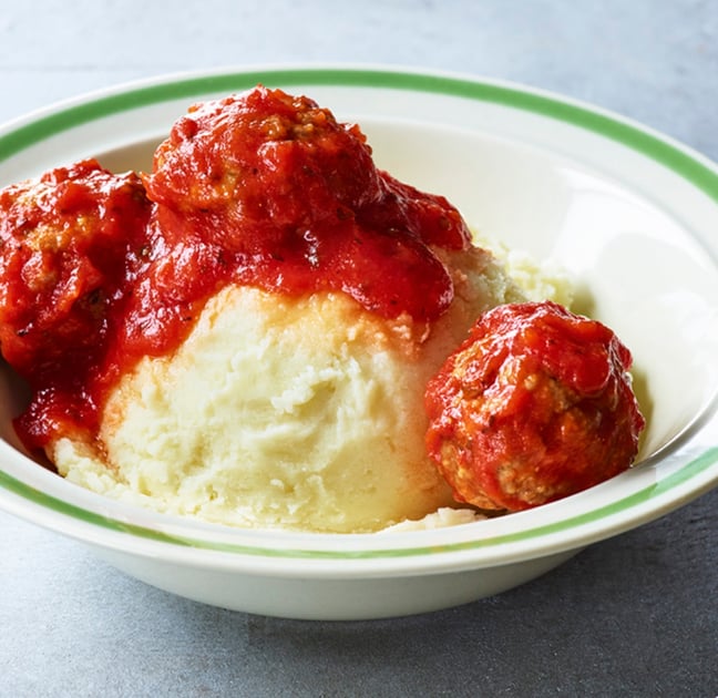 A bowl filled with mashed potatoes topped with three meatballs and tomato sauce, served in a white dish with a green-rimmed edge.