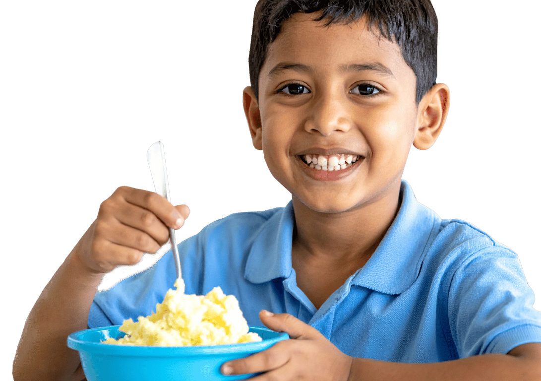 A smiling young boy in a blue shirt holds a blue bowl of mashed potatoes and a spoon.
