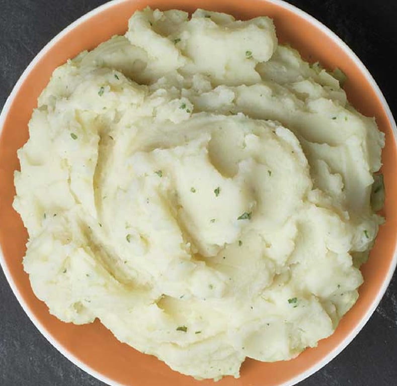 A bowl filled with creamy mashed potatoes, garnished with small flecks of green herbs, sits on a dark surface. The mashed potatoes appear smooth and fluffy.