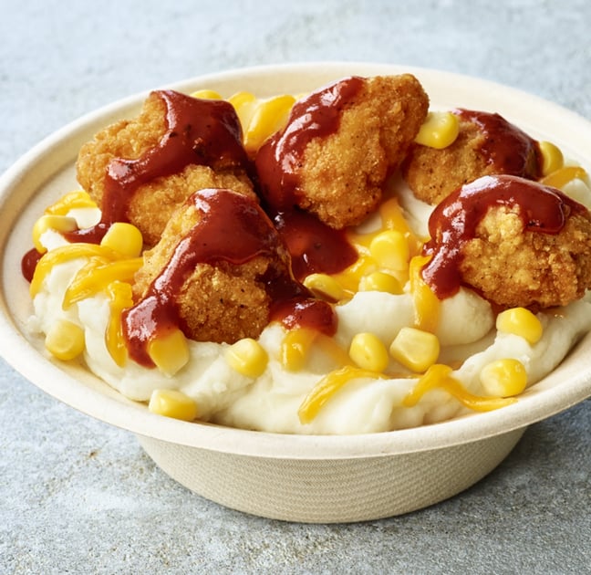 A bowl of mashed potatoes topped with corn, shredded cheese, crispy chicken pieces, and brown gravy.